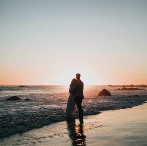 two people standing near the water on the beach while holding each other and watching the sunset