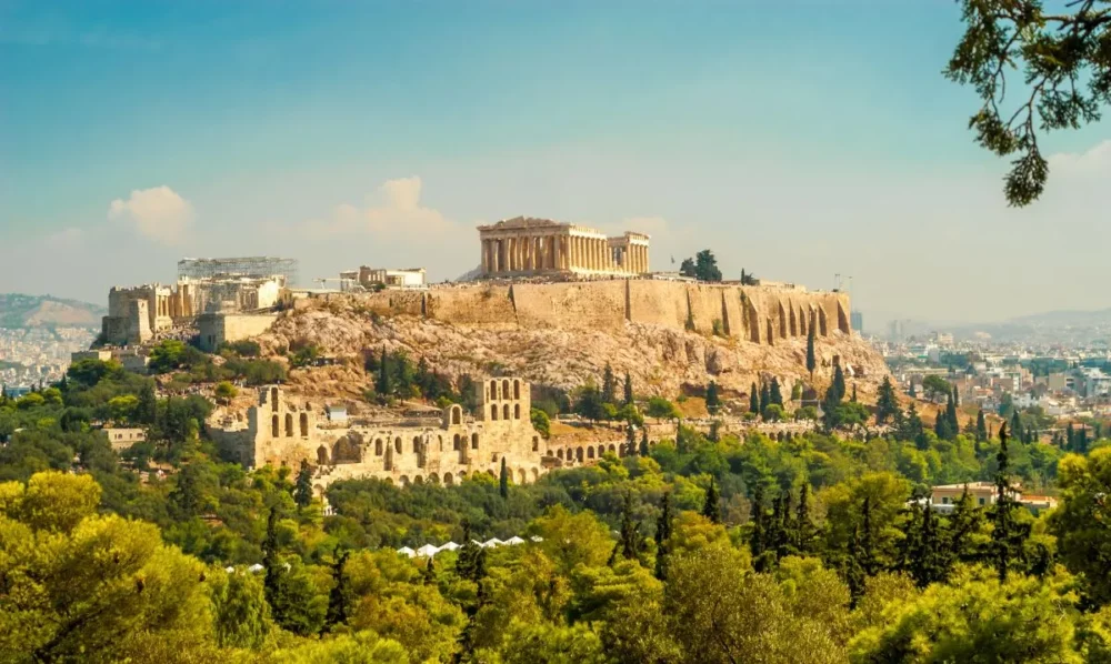 The Acropolis in Athens, a historic site with ancient ruins atop a hill, surrounded by lush greenery and the cityscape.