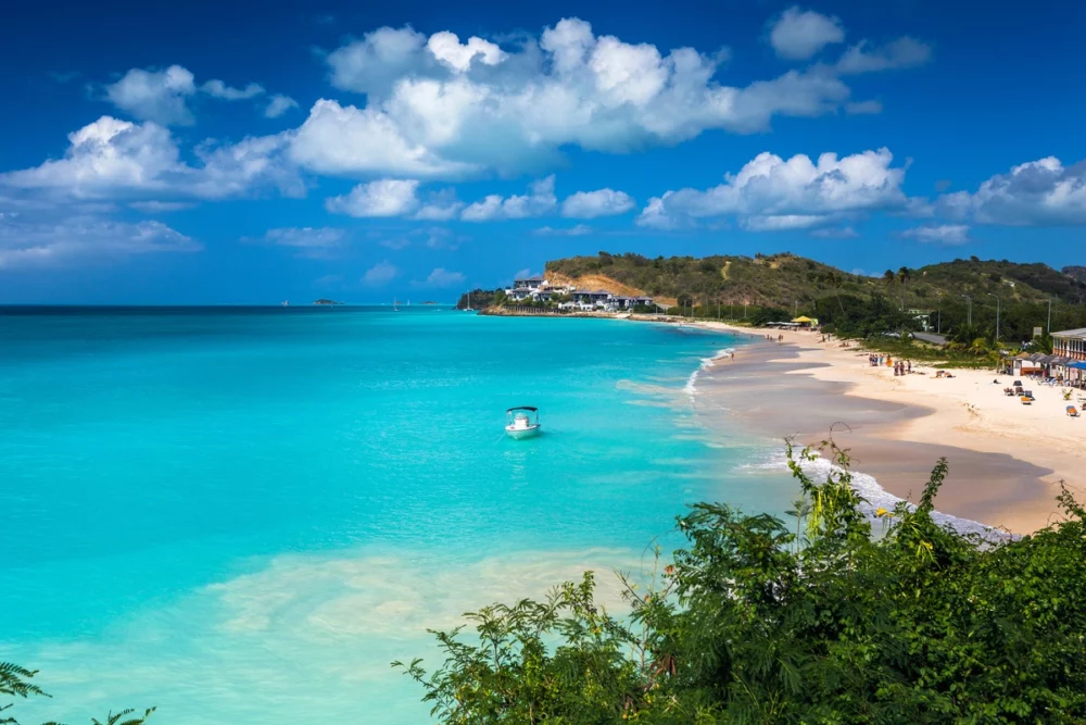 Relaxing scene of a docked boat on a beautiful beach in Antigua with turquoise water, white sand, and palm trees.