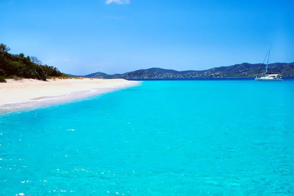 Buck Island near St. Croix, featuring a white sandy beach and turquoise waters, ideal for all-inclusive yacht charters, with a distant sailboat and hilly landscape in the background.






