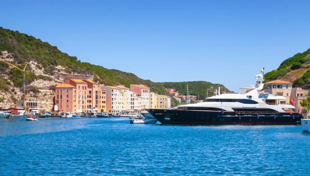  A large yacht docked in a harbor in Corsica, France. The yacht is surrounded by crystal-clear water and lush greenery.
