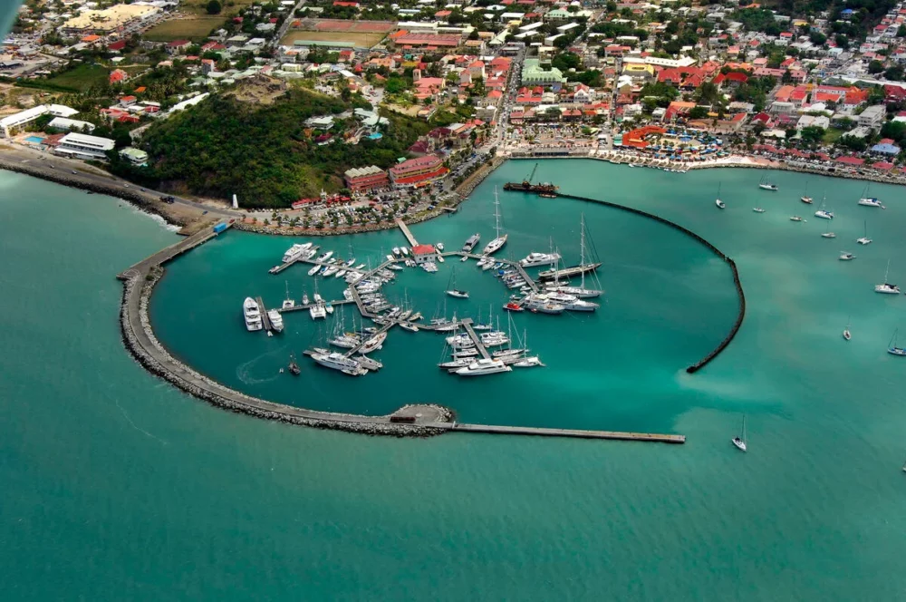 An aerial view of Fort Louis Marina on the Caribbean island of St. Martin, showcasing rows of yachts in crystal-clear turquoise water.