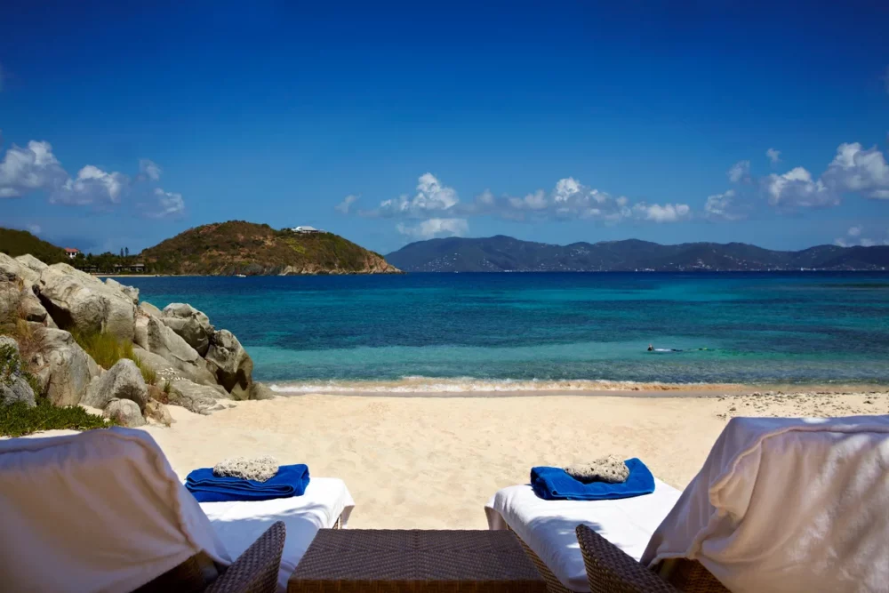 Empty lounge chairs sit on white sand overlooking turquoise water at Honeymoon Beach, British Virgin Islands, a private yacht rental Caribbean.
