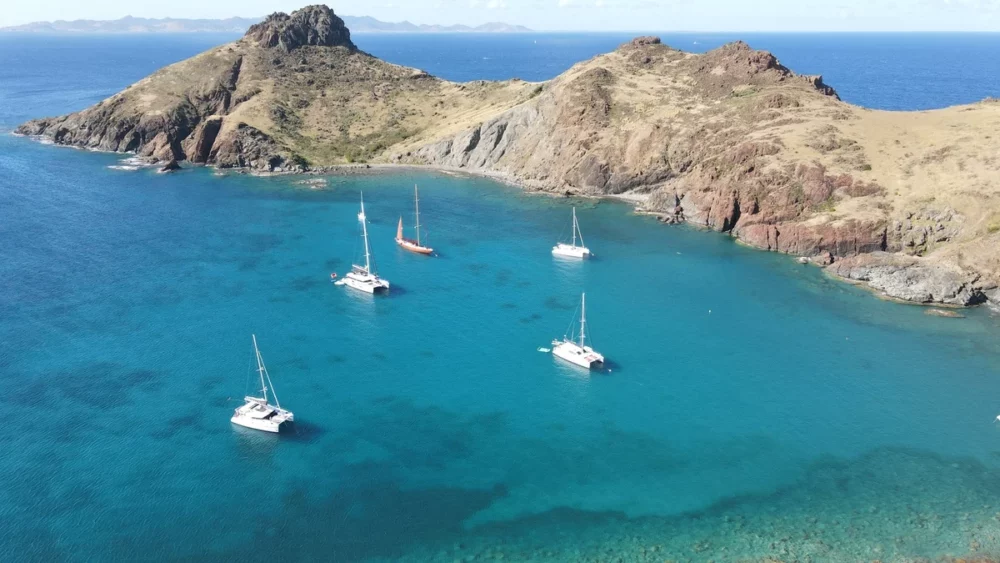 A group of sailboats docked in a calm bay next to a rocky island with palm trees. This image depicts the beauty of St. Barths and Ile Fourchue, a popular destination for private crewed yacht getaways.