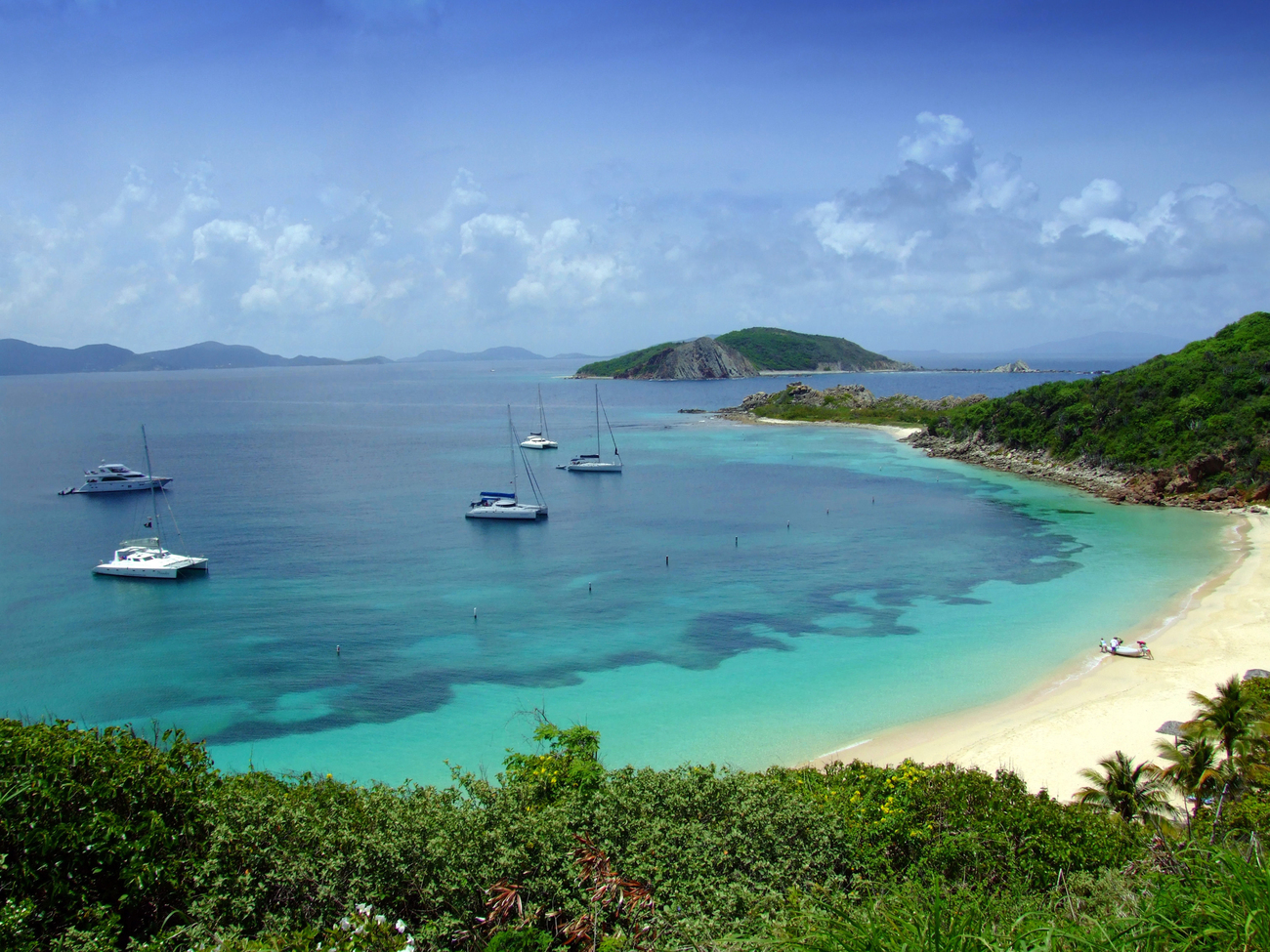 A tranquil view of Little Deadman's Beach with several yachts anchored in the clear turquoise waters, surrounded by lush greenery and distant island.