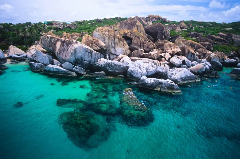 Aerial view of The Baths at Virgin Gorda, showcasing large granite boulders and clear turquoise waters, surrounded by lush greenery under a partly cloudy sky.
