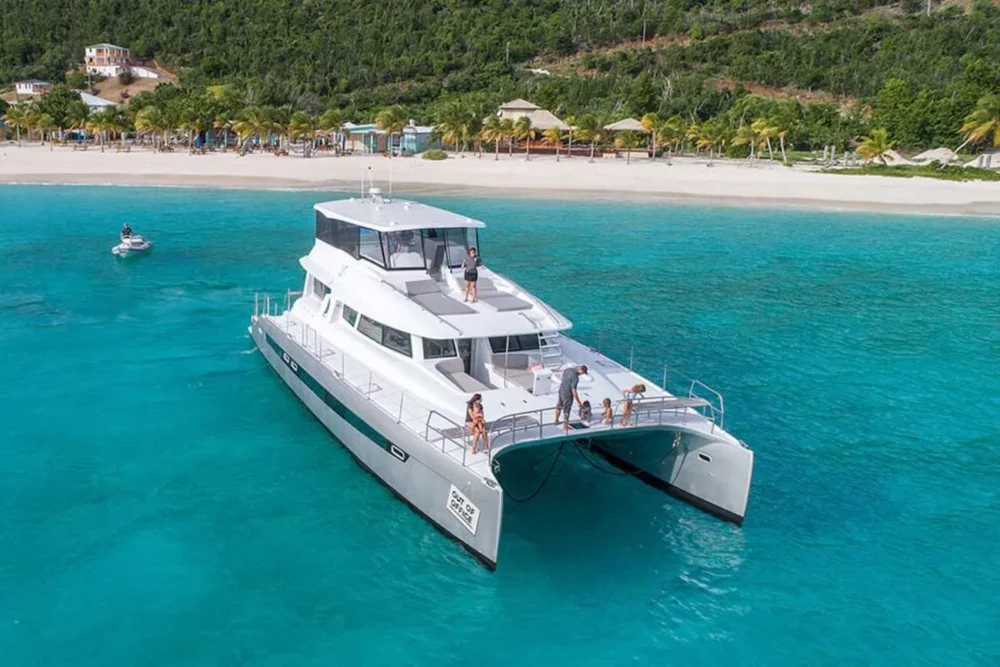 Voyage 650 Power Catamaran offering all-inclusive yacht charters, anchored in clear turquoise waters near a tropical beach with lush greenery and beachside houses.